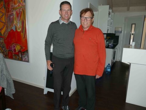 Zwei Männer lächeln in moderner Wohnküche; einer hält Mikrofon, Kunstbild im Hintergrund, Porträtfoto indoor