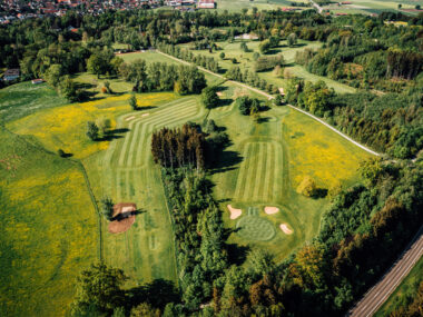Luftaufnahme Golfplatz in grüner Hügellandschaft mit Fairways, Greens und Bunkern nahe Landstraße