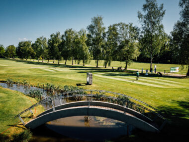 Sonniger Golfplatz mit Brücke über Teich, Golfer am Abschlag auf gepflegtem Fairway zwischen Birken.